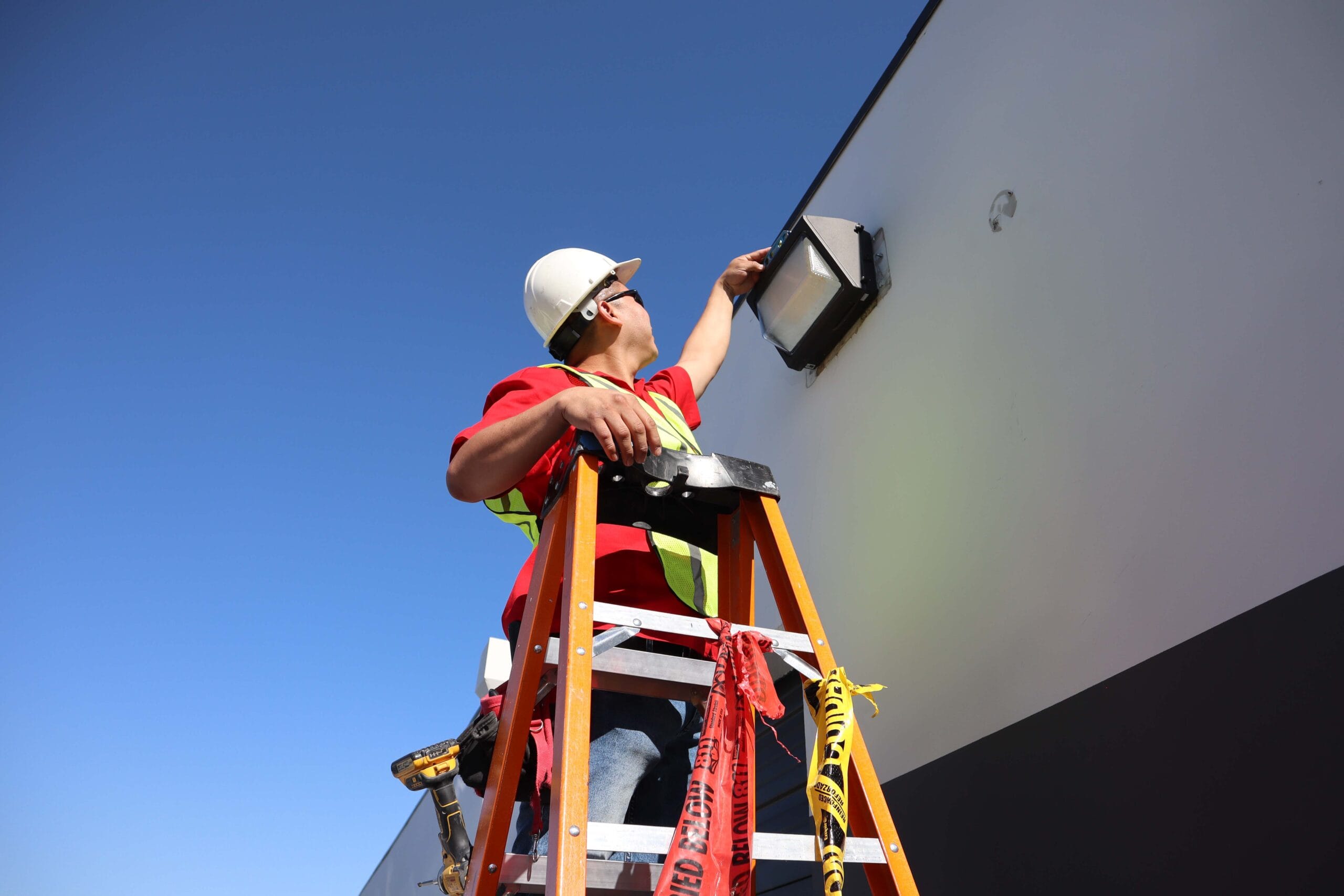 Licensed electrician on ladder installing outdoor lighting fixture on building exterior