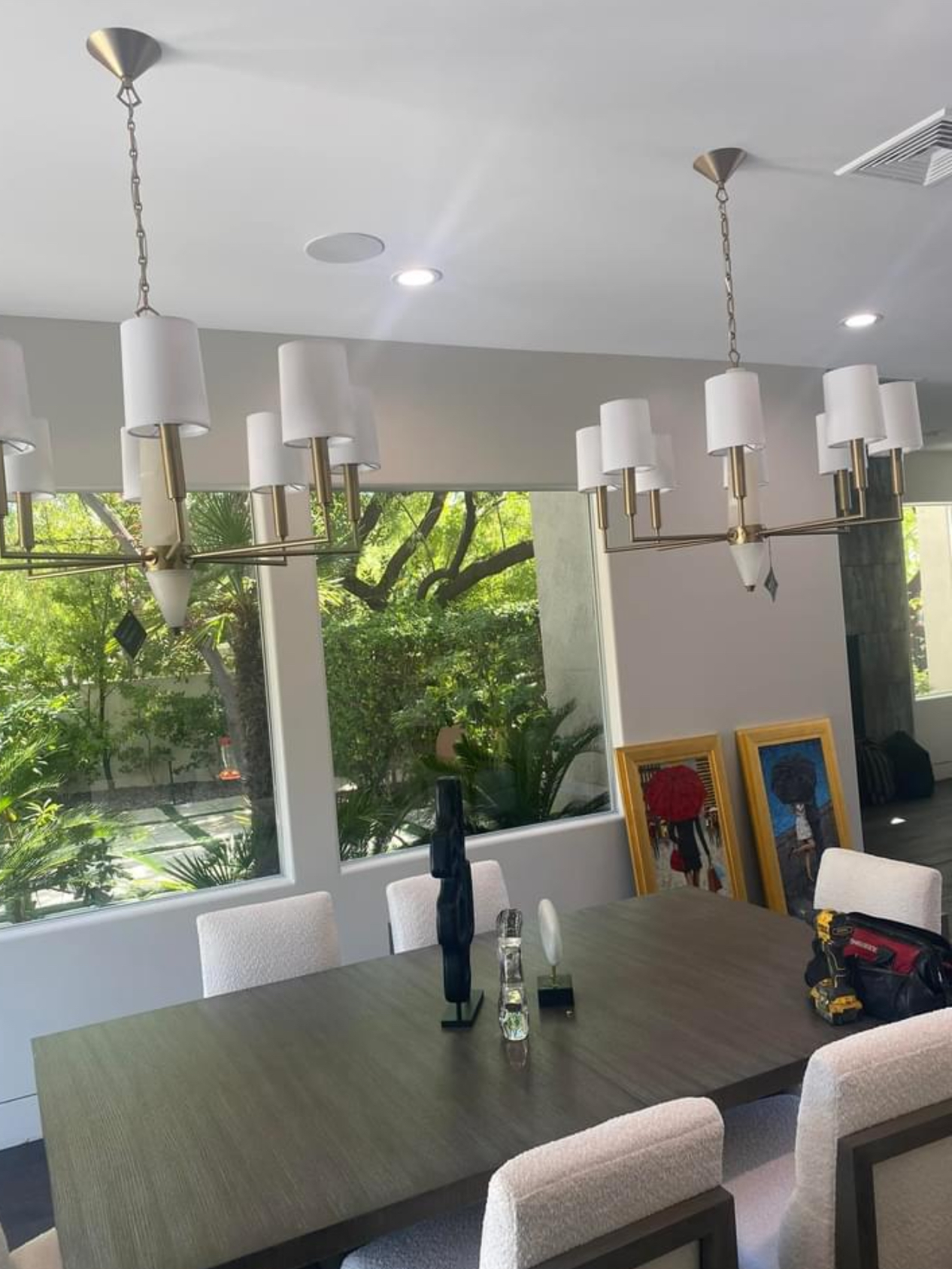 Modern dining room with a large dark wood table white upholstered chairs and brass chandeliers hanging from the ceiling large windows show greenery outside and framed artwork leaning against the wall inside
