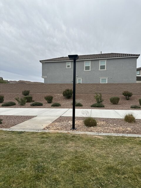 Black street light on a concrete sidewalk in a desert style front yard with a gray two story house and small shrubs in the background