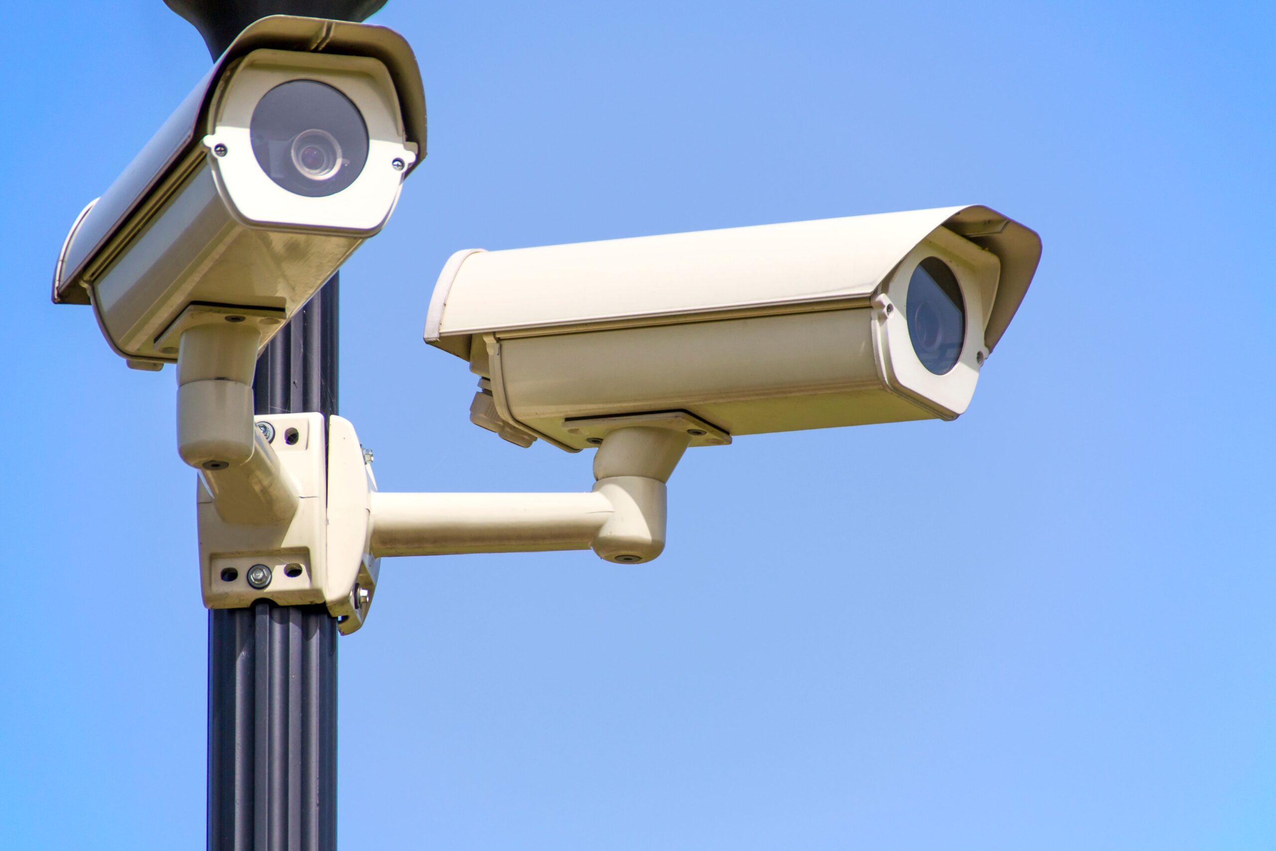 Two outdoor surveillance cameras mounted on a metal pole against a clear blue sky