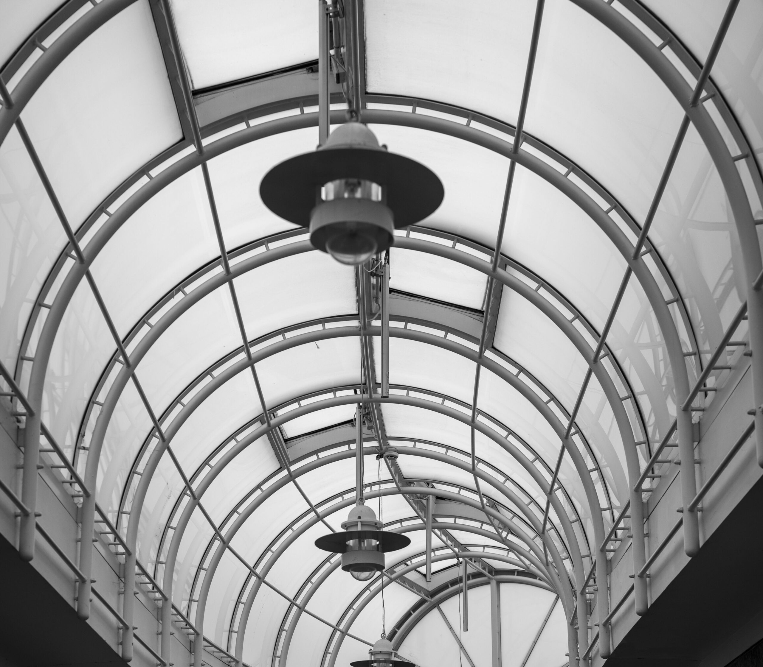 Black and white photo of curved metal arches forming a glass covered walkway with hanging lamps at the center